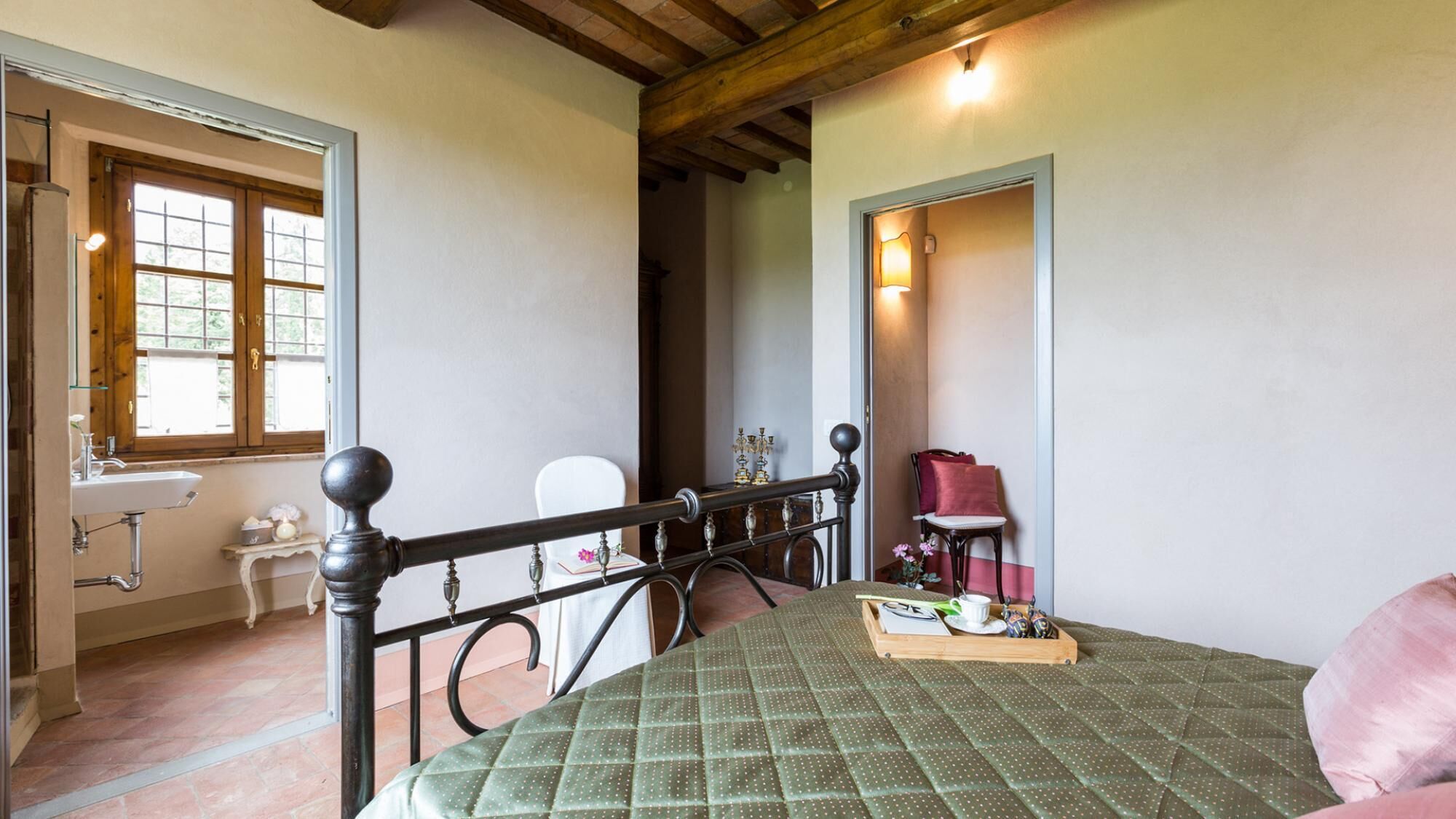 A bedroom with a bed, a chair, a small table with a tray, a window, and a bathroom visible in the background.