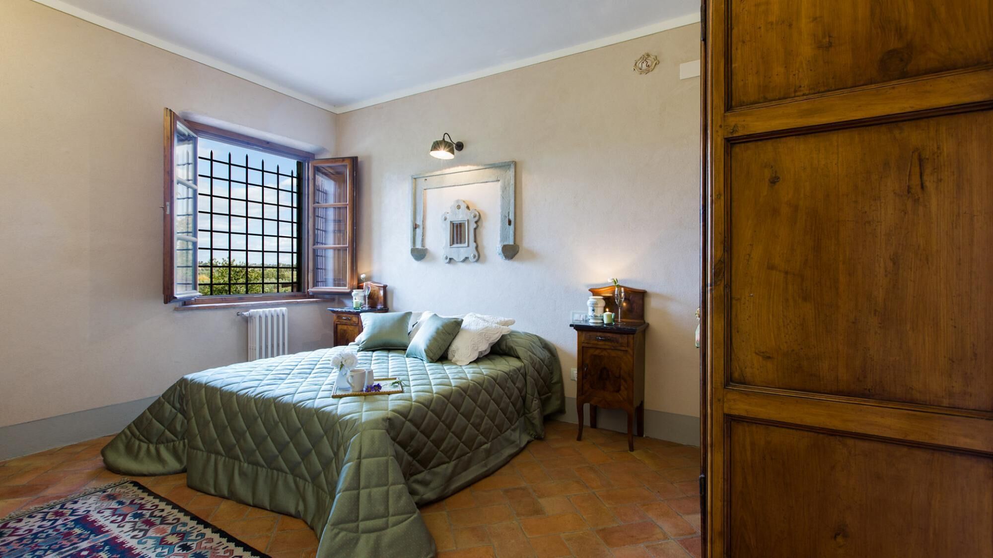 A bedroom with a bed, a window, a radiator, a wooden door, and a small bedside table with a lamp.