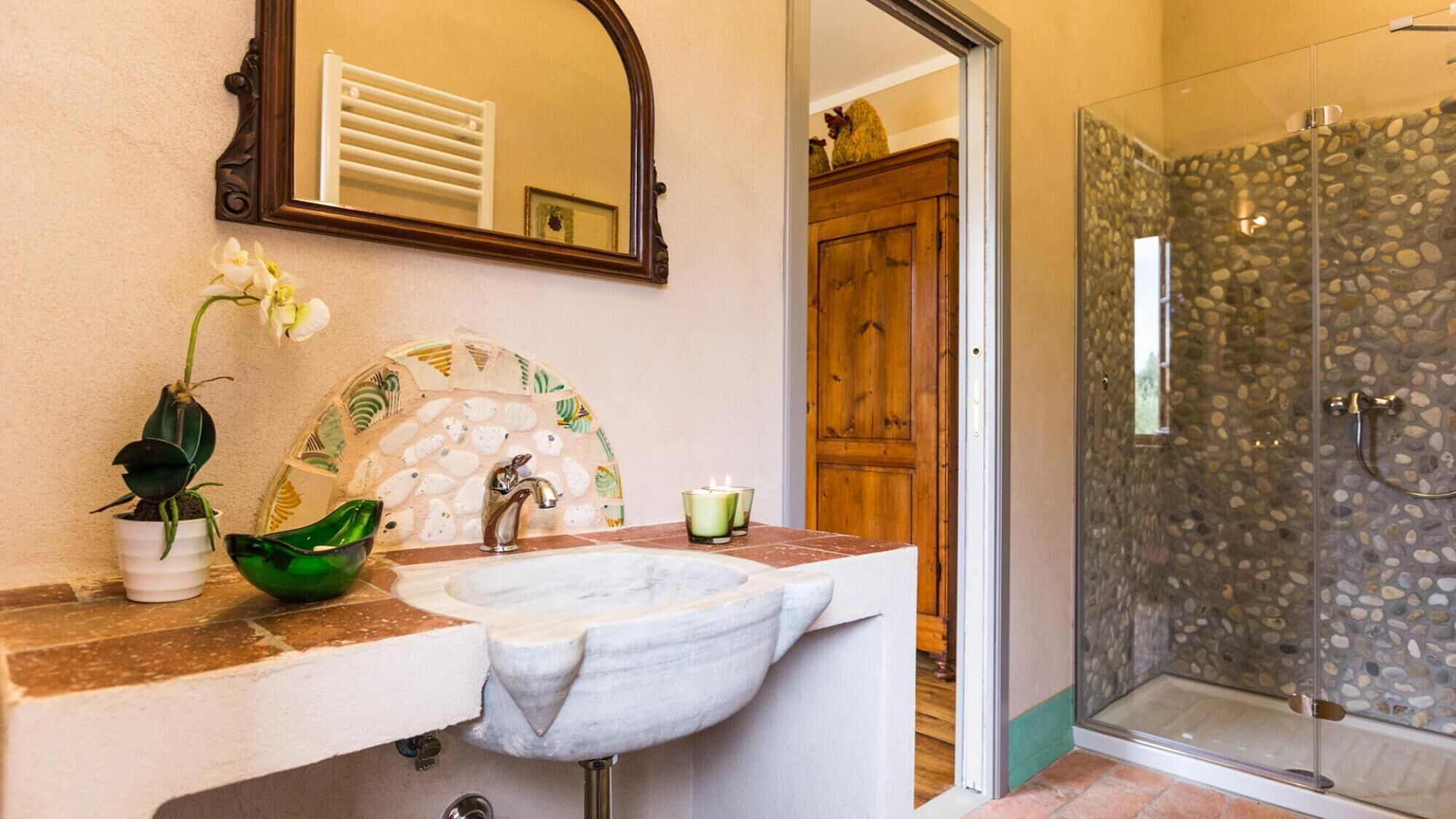 A bathroom with a marble sink, a mirror, a green bowl, and a shower with a stone wall.