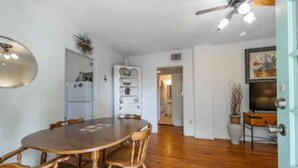 Un coin repas avec une table ronde en bois et des chaises en osier, un ventilateur de plafond et un réfrigérateur blanc en arrière-plan.
