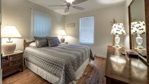 Une chambre à coucher comprenant un lit, des tables de chevet, une commode, un ventilateur de plafond et une fenêtre avec des stores.