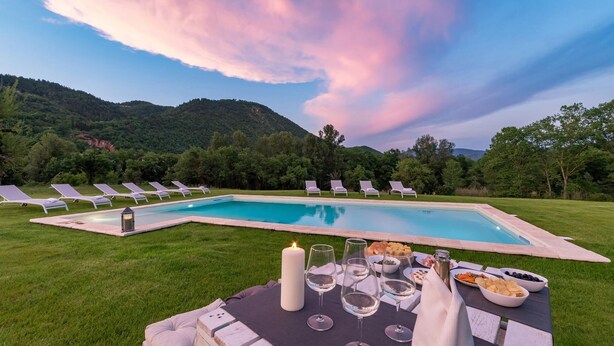 A poolside dining area with a table set for two, surrounded by lounge chairs, and a scenic view of mountains and trees. - Villa Monica 8 in Greti (Greve in Chianti)