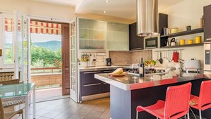 A modern kitchen with a large island, a dining area with red chairs, and a view of greenery through the open door. - Villa Monica 8 in Greti (Greve in Chianti)