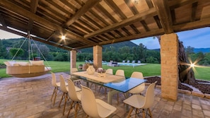 Uma área de refeições ao ar livre com mesa e cadeiras de vidro, uma cadeira suspensa e vista para uma piscina e vegetação.