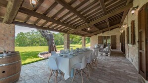 An outdoor covered patio with a long dining table set for a meal, stone walls, and a view of a grassy landscape. - Villa Monica 8 in Greti (Greve in Chianti)