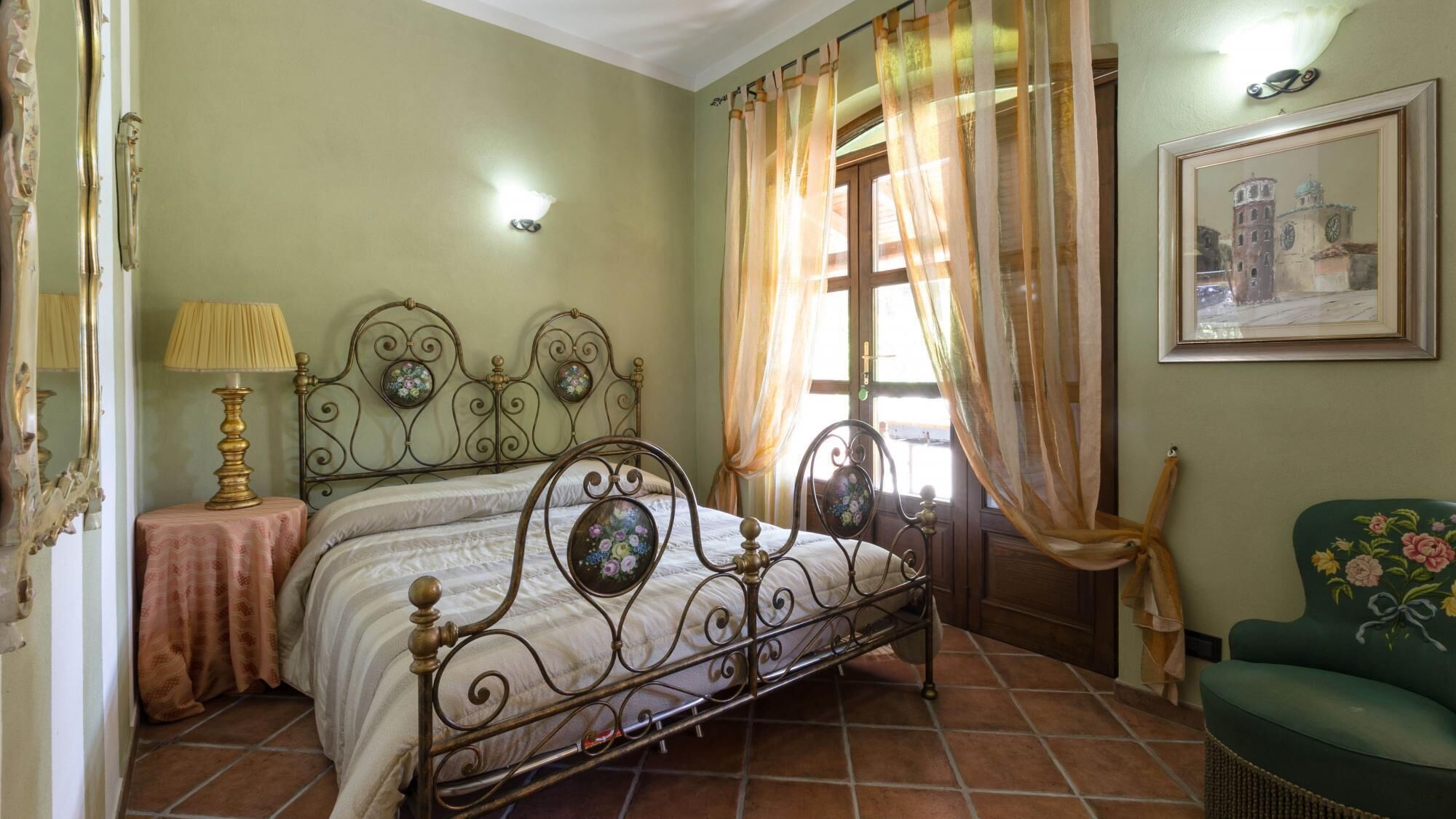 A bedroom with a metal bed frame, a bedside table with a lamp, a window with sheer curtains, and a framed picture on the wall.