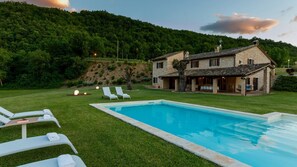 Une piscine avec des chaises longues, une maison en pierre dotée d’un patio et, en arrière-plan, une colline.