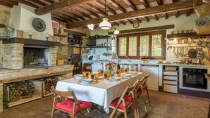 A rustic kitchen with a stone fireplace, wooden beams, and a dining table set for six with red cushioned chairs. - La Cerqua 8 in Gualdo Cattaneo (Gualdo Cattaneo)