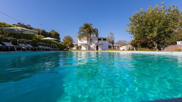 A clear blue pool with lounge chairs and umbrellas on the left, a white building with a covered entrance in the center, and trees on the right. - Villa D Aquino 8 2 in Casarano (Casarano)