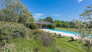 A pool with lounge chairs, a covered seating area, and lush greenery.