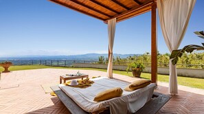 A covered outdoor seating area with a bed, a table, and a view of the landscape.