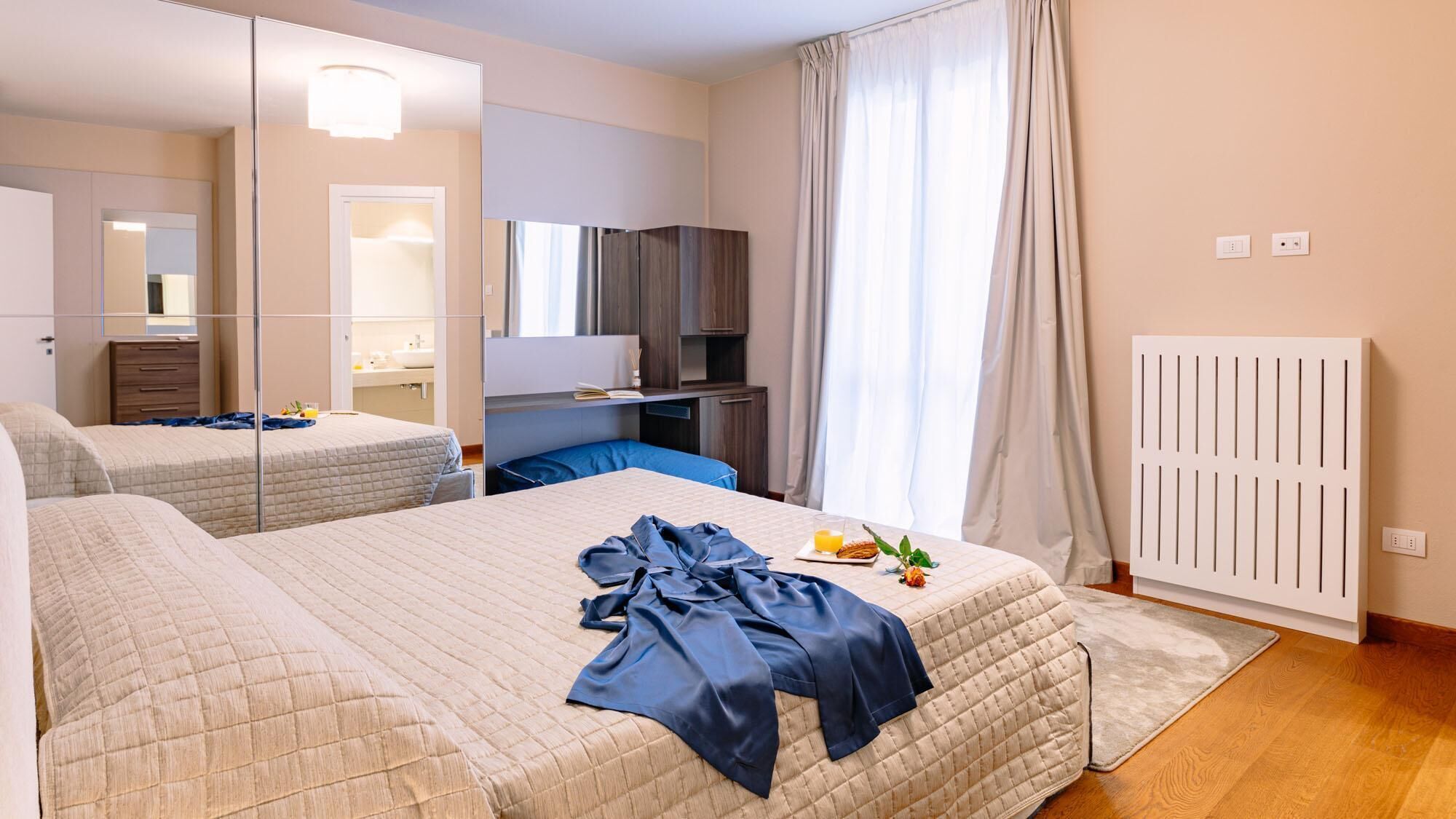 Une chambre d’hôtel avec deux lits, un bureau, un miroir, un radiateur et une fenêtre avec des rideaux.