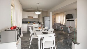 Uma cozinha moderna com armários brancos, uma sala de jantar com mesa posta para quatro pessoas e uma sala de estar com sofá e televisão.