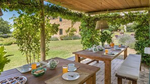 An outdoor dining area with wooden tables and benches, surrounded by greenery and a brick building in the background. - Casale Piattini 10 in Massa Bassa (Narni)