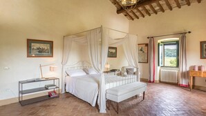 Quarto com cama de dossel, mesa de cabeceira, um banco e janela com vista para o verde.