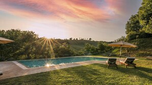 Piscina com deck pavimentado, cercada por grama e árvores. Há duas espreguiçadeiras com guarda-sóis.