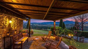 A stone patio with wooden chairs and a table, overlooking a scenic landscape with mountains and a sunset. - Casale del Viaggiatore 10 (San Casciano dei Bagni)