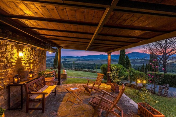 A stone patio with wooden chairs and a table, overlooking a scenic landscape with mountains and a sunset.