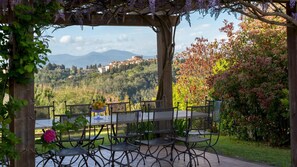 Un espacio de comedor al aire libre con vistas a un pueblo en la ladera, rodeado de vegetación y una pérgola.