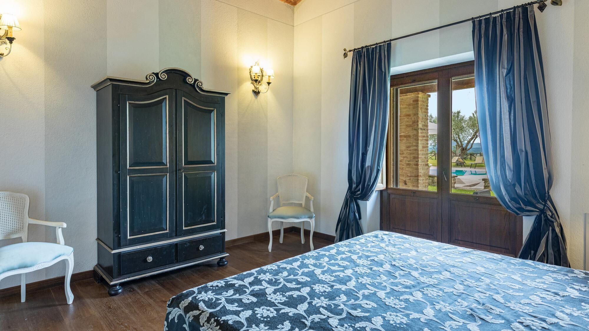A bedroom with a large bed, a wardrobe, two chairs, and a window with curtains.
