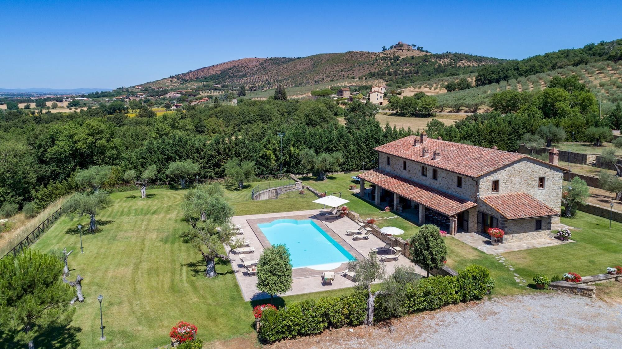 A large stone house with a swimming pool, surrounded by lush greenery and a hilly landscape.