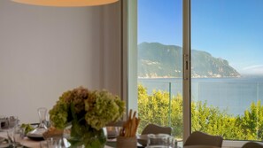 A dining area with a table set for two, a vase of flowers, and a view of the sea and mountains through large windows.
