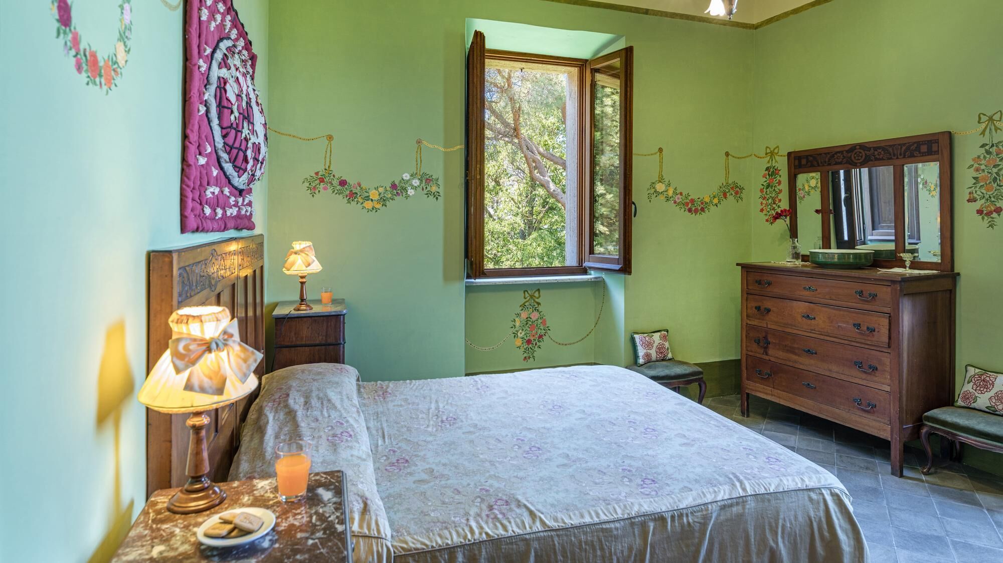 A bedroom with a bed, a dresser, a nightstand with a lamp, a window with a view of trees, and floral wall decorations.