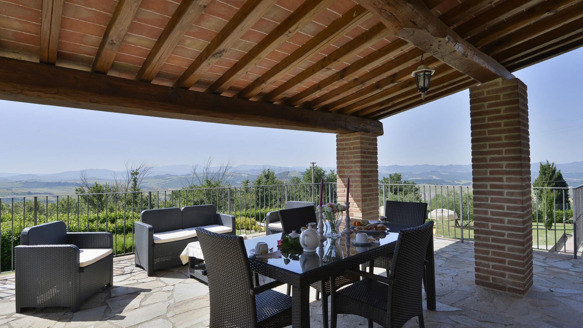 Patio esterno con mobili in vimini, un tavolo apparecchiato per mangiare e vista su montagne lontane.