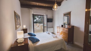 A bedroom with a bed, a dresser, a nightstand, a lamp, a mirror, and a door leading to an outdoor area with chairs and a table. - Tp-e974-ciav0at - Casale Dom 16 (Marsala)