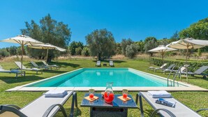Une piscine rectangulaire entourée de pelouse et d’arbres, avec des chaises longues et des parasols.