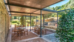 Une terrasse extérieure avec des tables et des chaises, un espace abrité doté d’un plafond en bois, et une vue sur un jardin et un mur en pierre.