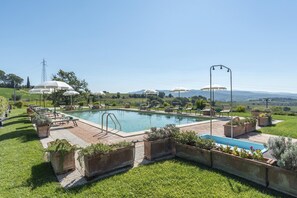A pool area with lounge chairs and umbrellas, surrounded by greenery and a view of distant hills.