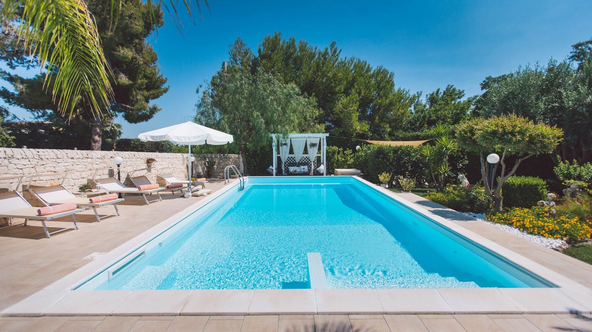 A rectangular swimming pool with a stone wall on one side and a white pergola with a canopy on the other.