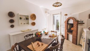 A dining area with a wooden table, chairs, and a variety of fruits and vegetables. There is a brick oven and a hanging light fixture above the table. - Rg-e366-sp460at - Villa Ferradina 4 2 (Ispica)