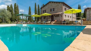 A swimming pool with lounge chairs and umbrellas, surrounded by stone buildings and tall trees.