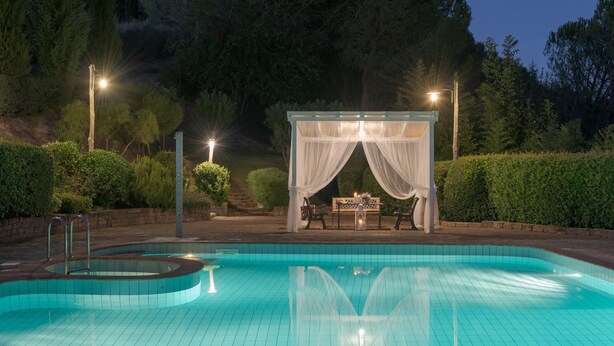 A pool area with a covered seating area and a table, surrounded by greenery and trees.