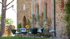 Un espace extérieur aménagé avec des sièges, un bâtiment en pierre en arrière-plan, une table et des chaises, et un arbre sur la gauche.
