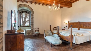 Une chambre avec une fenêtre en arc de pierre, un lit en bois, une chaise, un bureau et un lustre.