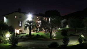 A two-story building with a well-lit entrance and a garden with various plants. - Lu-b648-capa0at - Villa Alida 12 (Capannori)