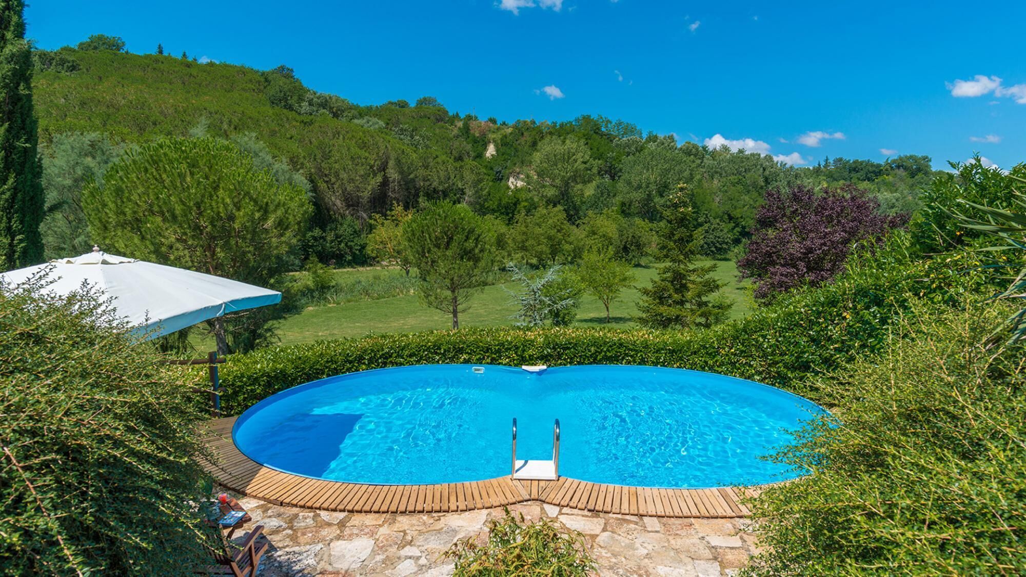 Une piscine dotée d’une terrasse en bois et d’un parasol blanc, entourée d’une végétation luxuriante et d’arbres.