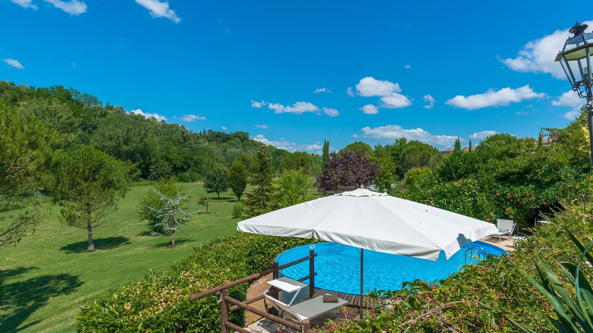 Una piscina con un ombrellone bianco, immersa nel verde e alberi.