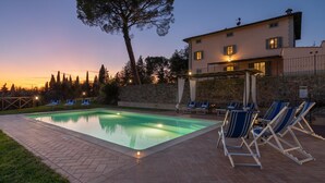 A swimming pool with lounge chairs, a stone wall, and a building with lights on in the background.
