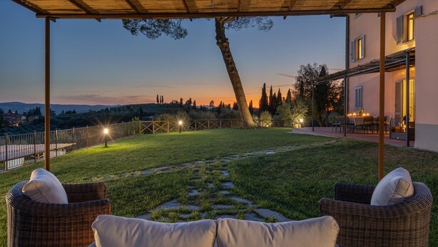 A covered outdoor area with wicker furniture, a grassy lawn, and a view of a hillside at dusk.