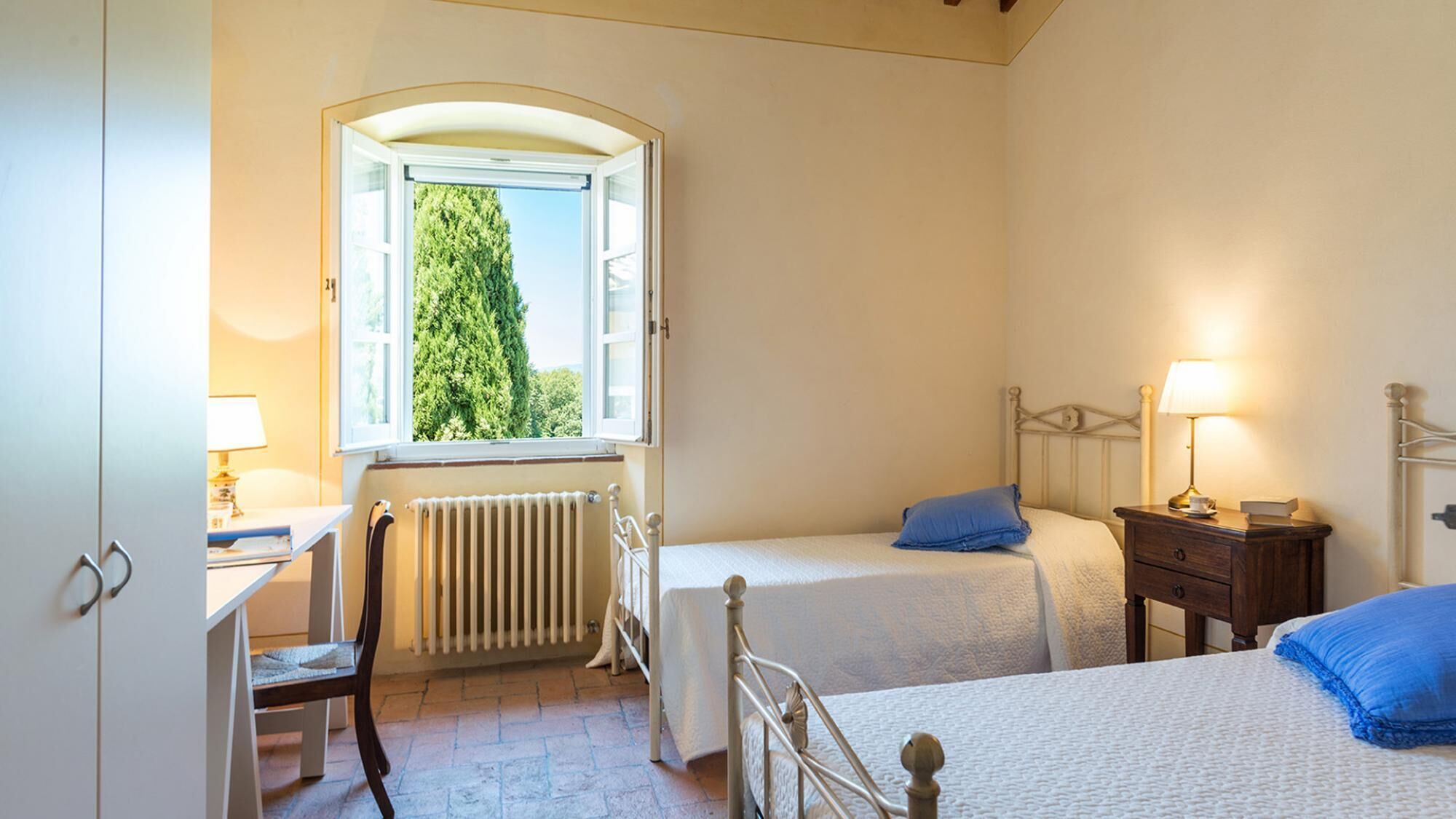 A bedroom with two beds, a radiator, a desk with a lamp, and a window with a view of trees.