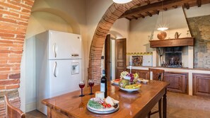 A rustic kitchen with a brick archway, a white refrigerator, and a wooden dining table set with food and wine. - Ar-f628-mssa0at - Villa Pucci 12 (Monte San Savino)