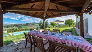 A covered outdoor dining area with a table set for four, overlooking a pool and scenic countryside.