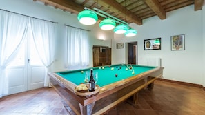 Uma mesa de bilhar com bolas e tacos, uma garrafa de cerveja e um copo sobre um tabuleiro. Três luminárias pendem sobre a mesa.