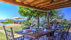 An outdoor dining area with a wooden table set for a meal, surrounded by chairs, under a wooden pergola.