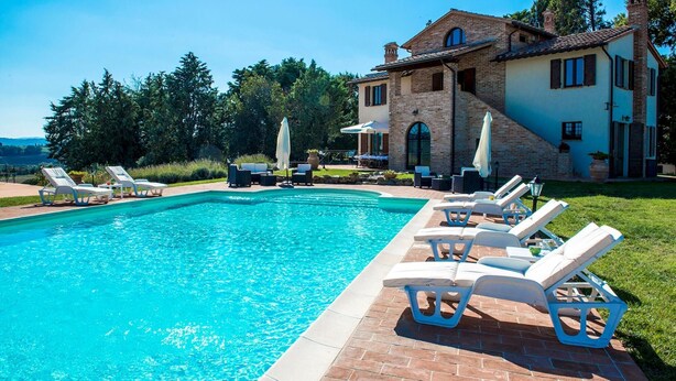 A swimming pool with lounge chairs and umbrellas, surrounded by a brick patio and a house with a tiled roof.