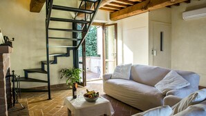 A cozy living room with a white sofa, a small table with a fruit bowl and wine glasses, a spiral staircase, and a view of the outdoors.